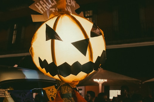 A large, illuminated jack-o'-lantern with a menacing smile and triangular eyes dominates the foreground. It appears to be part of a Halloween celebration, with the word 'HALLOWEEN' visible on a sign above. The background is dark and suggests a nighttime setting, possibly with people and decorations present.