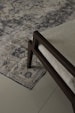 An ornate, patterned rug lies on a tiled floor. The rug's intricate design features various geometric and floral motifs in shades of blue and beige. A section of a modern chair with a dark wooden frame and light-colored cushion is visible on the rug.