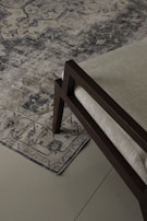 An ornate, patterned rug lies on a tiled floor. The rug's intricate design features various geometric and floral motifs in shades of blue and beige. A section of a modern chair with a dark wooden frame and light-colored cushion is visible on the rug.