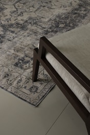 An ornate, patterned rug lies on a tiled floor. The rug's intricate design features various geometric and floral motifs in shades of blue and beige. A section of a modern chair with a dark wooden frame and light-colored cushion is visible on the rug.