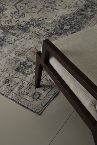 An ornate, patterned rug lies on a tiled floor. The rug's intricate design features various geometric and floral motifs in shades of blue and beige. A section of a modern chair with a dark wooden frame and light-colored cushion is visible on the rug.