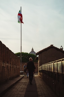 A person walks down a narrow cobblestone pathway lined with brick walls on a clear day. An Italian flag waves on a flagpole, and in the background, there is a domed building partially obscured by trees.