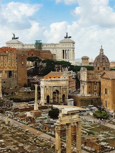 Ancient Roman ruins are spread across a vast open area. Large columns and remnants of historical structures dominate the foreground, while a grand, ornate building with classical architecture stands in the background. Sculptures and statues adorn the top of the building, and there's a large archway amongst the ruins. The sky above features fluffy white clouds against a blue backdrop.