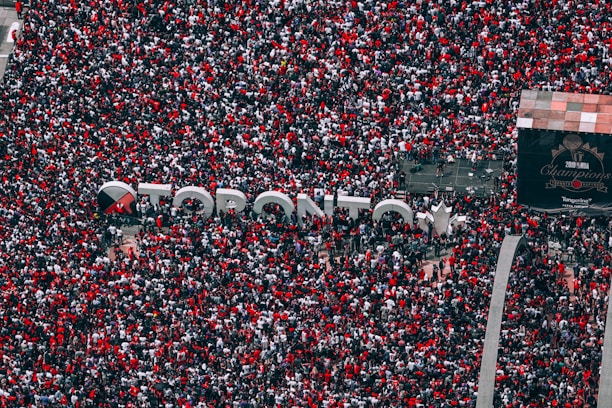 A massive crowd gathers around a large sign spelling 'Toronto' during a celebration. People are predominantly wearing red, interspersed with patches of white and black clothing. The crowd fills the image from edge to edge, and a large screen in the background references a 2019 basketball championship.