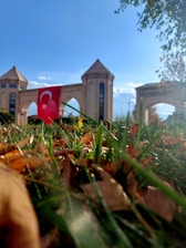 A monument or entrance with three towers and a large archway is seen in the background. A Turkish flag is prominently displayed in front of the structure. The foreground is dominated by green grass and dried brown leaves, with clear blue skies and some clouds overhead.