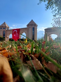 A monument or entrance with three towers and a large archway is seen in the background. A Turkish flag is prominently displayed in front of the structure. The foreground is dominated by green grass and dried brown leaves, with clear blue skies and some clouds overhead.