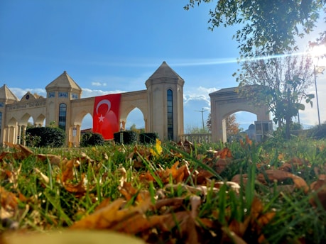 A grand architectural entrance with pointed arches and towers stands prominently. The building displays a large red flag with a crescent moon and star. In the foreground, vibrant green grass and scattered autumn leaves are visible, with some trees and bushes in the background under a clear blue sky.
