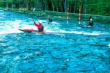 Several kayakers navigate turbulent waters in an outdoor setting. The scene is vibrant with lush greenery surrounding the river. The kayakers wear helmets and life vests as they maneuver through the water, using their paddles to control their movement. Red and white poles are positioned along the course for navigation.
