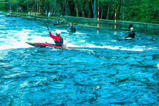Several kayakers navigate turbulent waters in an outdoor setting. The scene is vibrant with lush greenery surrounding the river. The kayakers wear helmets and life vests as they maneuver through the water, using their paddles to control their movement. Red and white poles are positioned along the course for navigation.
