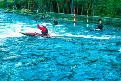 Several kayakers navigate turbulent waters in an outdoor setting. The scene is vibrant with lush greenery surrounding the river. The kayakers wear helmets and life vests as they maneuver through the water, using their paddles to control their movement. Red and white poles are positioned along the course for navigation.