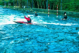 Several kayakers navigate turbulent waters in an outdoor setting. The scene is vibrant with lush greenery surrounding the river. The kayakers wear helmets and life vests as they maneuver through the water, using their paddles to control their movement. Red and white poles are positioned along the course for navigation.