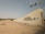 A desert landscape with a prominent sign indicating a luxury collection desert resort and spa called Al Maha. The sign is mounted on a textured stone wall with several poles holding flags above it. The surrounding area features sandy terrain with sparse desert vegetation.