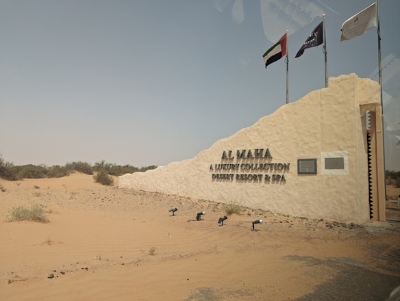 A desert landscape with a prominent sign indicating a luxury collection desert resort and spa called Al Maha. The sign is mounted on a textured stone wall with several poles holding flags above it. The surrounding area features sandy terrain with sparse desert vegetation.