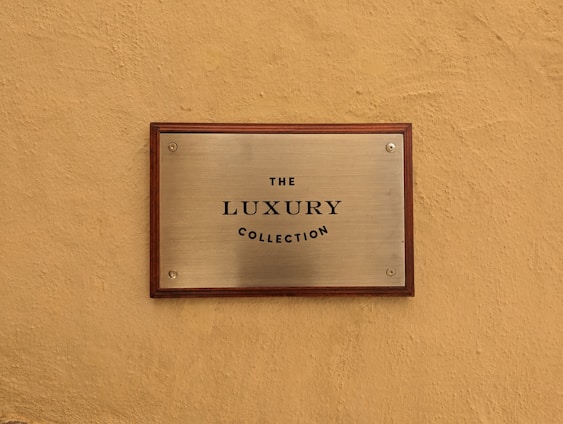 A metallic plaque with a wooden frame mounted on a textured beige wall. The plaque has the text 'The Luxury Collection' engraved in black, centered on its surface.