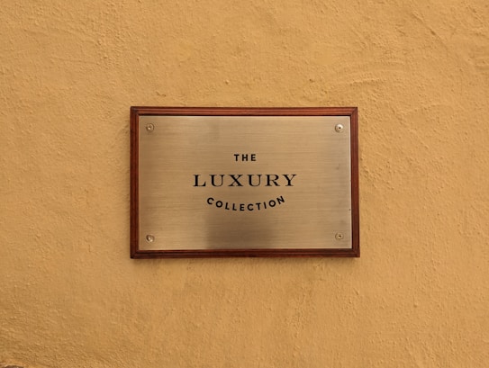 A metallic plaque with a wooden frame mounted on a textured beige wall. The plaque has the text 'The Luxury Collection' engraved in black, centered on its surface.