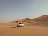 A four-wheel-drive vehicle is parked in a vast desert landscape with rolling sand dunes and a clear sky. A solitary tree stands near the dunes, adding contrast to the sandy environment.