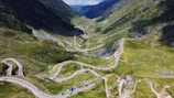 A scenic mountain landscape featuring a winding road that snakily curves through lush green hills. The road is lined with small, scattered vehicles. The backdrop showcases high, verdant peaks under a bright blue sky.