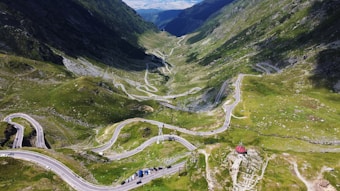 A scenic mountain landscape featuring a winding road that snakily curves through lush green hills. The road is lined with small, scattered vehicles. The backdrop showcases high, verdant peaks under a bright blue sky.