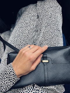 A person wearing a black and white leopard print outfit, holding a black leather clutch purse. The hand visible has a ring on one of the fingers.