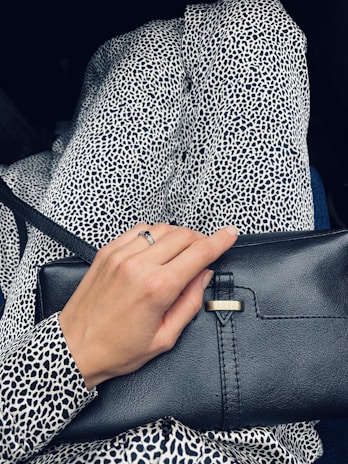 A person wearing a black and white leopard print outfit, holding a black leather clutch purse. The hand visible has a ring on one of the fingers.