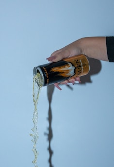 A hand is holding a can of energy drink, pouring its contents onto a surface. The liquid is captured mid-flow with a smooth stream.