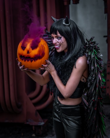 A person dressed in a dark, gothic costume holds a carved pumpkin with purple smoke emanating from it. The person is wearing black leather pants, a black top, and a fur-like cape. They have horns on their head and dramatic makeup with face paint that adds to the Halloween theme.