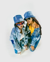 Two individuals are posing together while dressed in stylish denim outfits. Both are wearing bucket hats and sunglasses. One person is wearing a yellow top with a denim jacket and pants, while the other is wearing a white top, a distressed denim jacket, and denim pants.