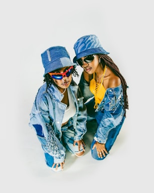 Two individuals are posing together while dressed in stylish denim outfits. Both are wearing bucket hats and sunglasses. One person is wearing a yellow top with a denim jacket and pants, while the other is wearing a white top, a distressed denim jacket, and denim pants.