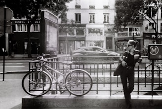A black and white urban scene features a woman leaning against a metal railing near a bicycle. She is wearing dark sunglasses and a leather jacket, exuding a casual and relaxed attitude. The background shows a street with a moving car, several buildings with shopfronts, and trees partially obscuring them.