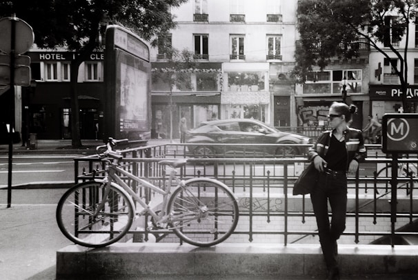 A black and white urban scene features a woman leaning against a metal railing near a bicycle. She is wearing dark sunglasses and a leather jacket, exuding a casual and relaxed attitude. The background shows a street with a moving car, several buildings with shopfronts, and trees partially obscuring them.