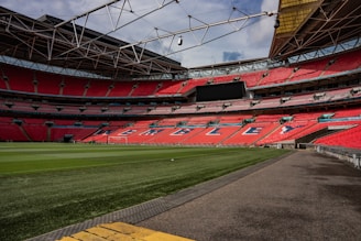 An empty football stadium with red seats, a lush green field, and a clear sky overhead. The stadium features a modern design with a spacious seating arrangement and a large electronic scoreboard.