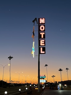 A tall, vintage-style motel sign is prominently displayed against a twilight sky, featuring illuminated letters spelling 'MOTEL.' The sign includes neon outlines of two people, one in red and the other in blue, depicted as if one is diving towards the ground. Palm trees line the road leading to the motel, with streetlights casting soft glows. The background sky transitions smoothly from deep blue to a warm orange near the horizon, capturing the tranquil, end-of-day ambiance.