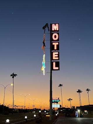 A tall, vintage-style motel sign is prominently displayed against a twilight sky, featuring illuminated letters spelling 'MOTEL.' The sign includes neon outlines of two people, one in red and the other in blue, depicted as if one is diving towards the ground. Palm trees line the road leading to the motel, with streetlights casting soft glows. The background sky transitions smoothly from deep blue to a warm orange near the horizon, capturing the tranquil, end-of-day ambiance.