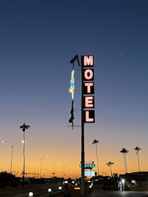A vintage motel sign illuminated with neon lights stands tall against a twilight sky. The word 'Motel' is prominently displayed vertically in bright red letters. A neon figure of a diver hangs beside the sign. Palm trees line the street in the foreground, and the sky transitions from a warm orange near the horizon to a deep blue higher up.