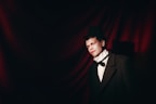 A person dressed in formal vintage attire, featuring a classic black top hat, white shirt with a bow tie, and an overcoat. The background consists of dark red fabric draped in soft waves, creating a dramatic and theatrical atmosphere.