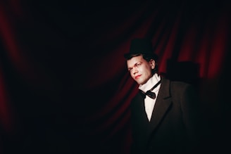 A person dressed in formal vintage attire, featuring a classic black top hat, white shirt with a bow tie, and an overcoat. The background consists of dark red fabric draped in soft waves, creating a dramatic and theatrical atmosphere.