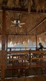 A rustic beachfront bar with wooden furniture and a thatched roof. The interior is shaded and features a ceiling fan. A person is sitting at one of the tables, facing the ocean view in the background.