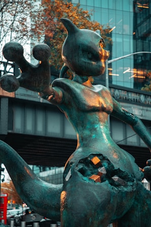 A large sculpture of a fictional creature stands in an urban setting, capturing attention with its dynamic pose. The sculpture has a metallic surface with a patina finish, and features geometric shapes embedded into the lower section. In the background, there are modern glass buildings and autumn trees, adding contrast to the artwork.