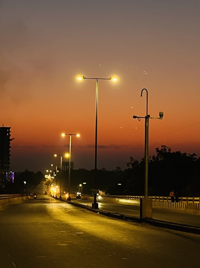 A road stretches into the distance under a dusky sky transitioning from warm oranges to deeper blues. Streetlights cast a yellow glow onto the asphalt, guiding the way. Silhouettes of trees and a building line the sides of the road. The atmosphere is tranquil, emphasizing solitude and calmness.