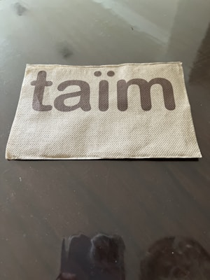 A fabric or textured item with the word 'taïm' printed on it in bold, brown letters. The surface is placed against a dark reflectively polished background.