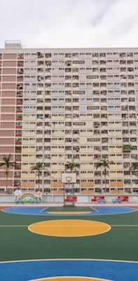 A high-rise residential building with a colorful facade featuring numerous windows and balconies. In front of the building, there is a vibrant basketball court with bright circles painted on the ground, surrounded by palm trees. The scene includes a small mural with cartoon characters and a rainbow.