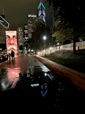 A nighttime cityscape featuring a large digital display with a close-up image of a person's face. The scene includes modern skyscrapers illuminated with lights, one of which displays the name 'Prudential'. The pavement is wet, reflecting the lights and the digital display. Several people are walking along the path under streetlights, surrounded by trees.
