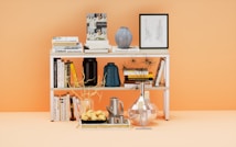 A neatly arranged white bookshelf stands against a peach-colored background. It is filled with various books, including titles about architecture, design, and literature. On the top shelf, there is a blue vase and a framed black-and-white sketch. The middle shelf holds two dark vases and a couple of abstract sculptures or decorative branches. The bottom shelf has more books and a tray with croissants beside a modern silver coffee pot and glass containers.
