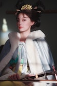 A person wearing traditional clothing, with a luxurious fur-lined cape and ornate headdress featuring gold and jewels. The attire includes layers of pale blue, green, and yellow fabric. The scene is softly lit, creating elegant shadows.
