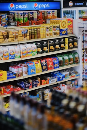 A well-organized supermarket shelf displaying a variety of snacks including cookies, crackers, and confectionery along with a prominent Pepsi logo at the top. The shelves are stocked with items like Milano cookies, Goldfish, Pringles, and Honey Bunches of Oats. The products are colorful, and the display is neatly arranged.