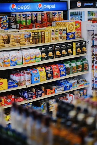 A well-organized supermarket shelf displaying a variety of snacks including cookies, crackers, and confectionery along with a prominent Pepsi logo at the top. The shelves are stocked with items like Milano cookies, Goldfish, Pringles, and Honey Bunches of Oats. The products are colorful, and the display is neatly arranged.