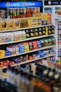 A well-organized supermarket shelf displaying a variety of snacks including cookies, crackers, and confectionery along with a prominent Pepsi logo at the top. The shelves are stocked with items like Milano cookies, Goldfish, Pringles, and Honey Bunches of Oats. The products are colorful, and the display is neatly arranged.