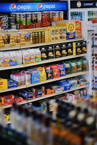 A well-organized supermarket shelf displaying a variety of snacks including cookies, crackers, and confectionery along with a prominent Pepsi logo at the top. The shelves are stocked with items like Milano cookies, Goldfish, Pringles, and Honey Bunches of Oats. The products are colorful, and the display is neatly arranged.