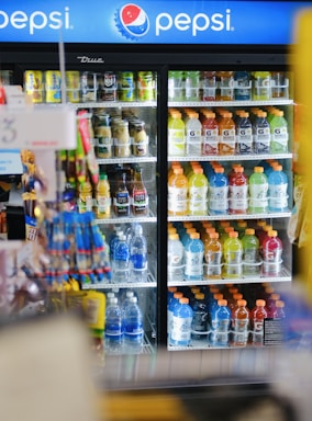 A refrigerated display case is filled with various beverages, including Gatorade, Aquafina water bottles, and other drinks in bottles and cans. The door of the cooler is made of clear glass, allowing a view of the colorful drinks arranged methodically inside. In the foreground, some snacks or items are slightly blurred. Above, a Pepsi sign is prominently visible, indicating the type of products stored within.