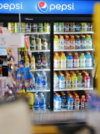 A refrigerated display case is filled with various beverages, including Gatorade, Aquafina water bottles, and other drinks in bottles and cans. The door of the cooler is made of clear glass, allowing a view of the colorful drinks arranged methodically inside. In the foreground, some snacks or items are slightly blurred. Above, a Pepsi sign is prominently visible, indicating the type of products stored within.